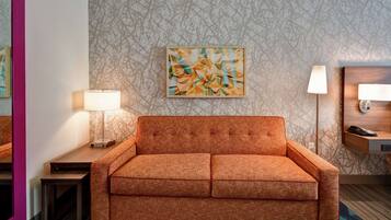Una habitación de hotel moderna con sofá, una mesita auxiliar con lámpara, una obra de arte colgada en la pared y una mesita de noche de madera con lámpara.