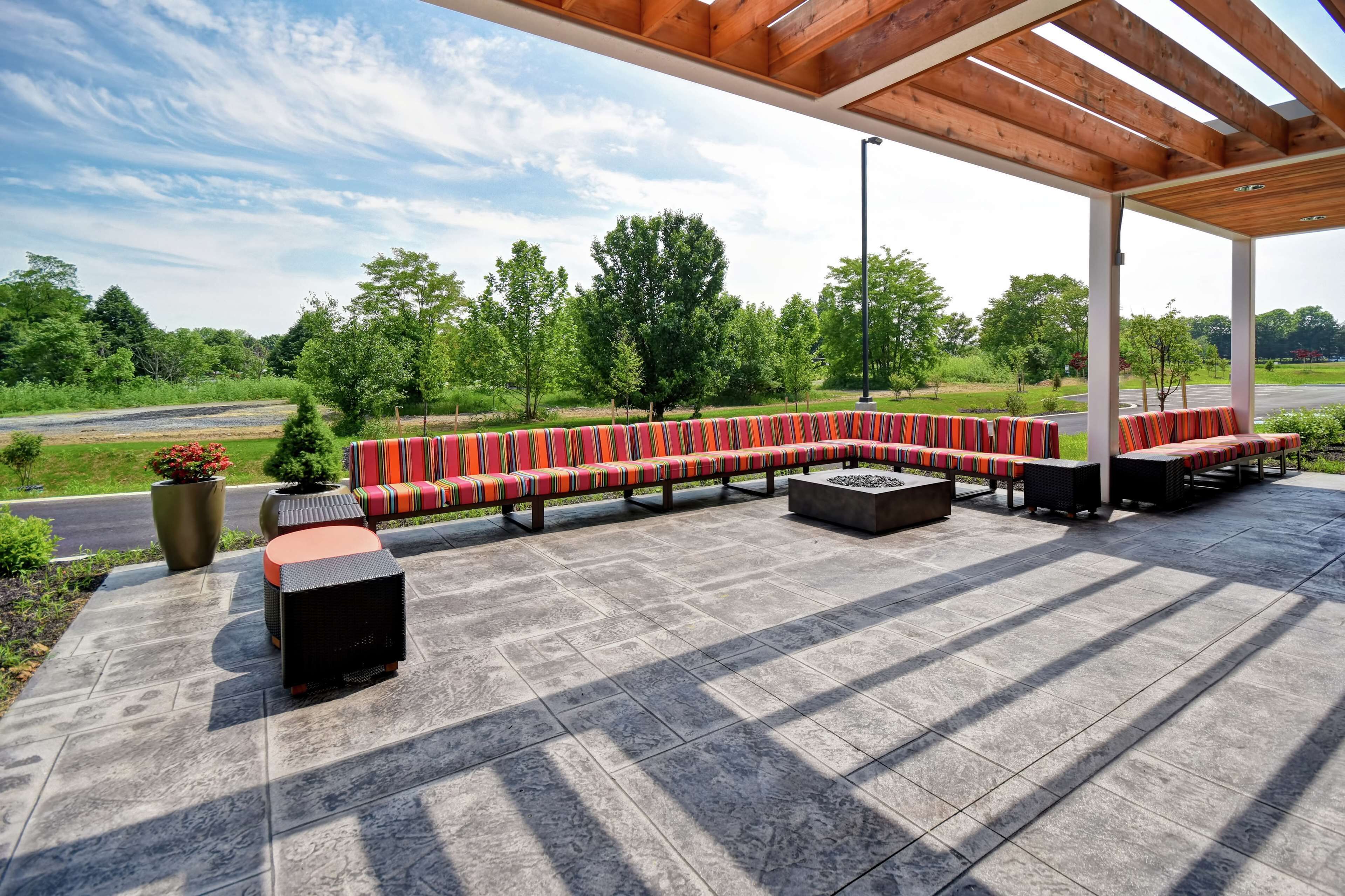 A covered outdoor seating area with a long bench and small tables, surrounded by a paved patio.