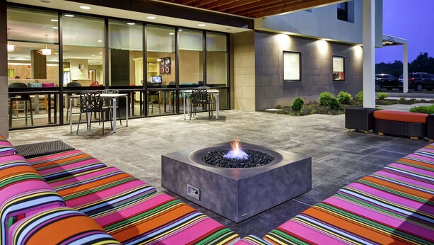 An outdoor seating area with colorful striped cushions, a fire pit, and a modern building with large windows and a bar area visible inside. - Home2 Suites by Hilton Mechanicsburg (Mechanicsburg)