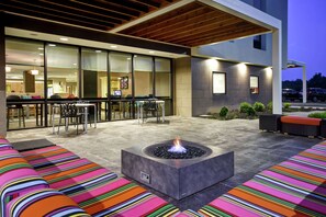 An outdoor seating area with colorful striped cushions, a fire pit, and a modern building with large windows and a bar area visible inside. - Home2 Suites by Hilton Mechanicsburg (Mechanicsburg)