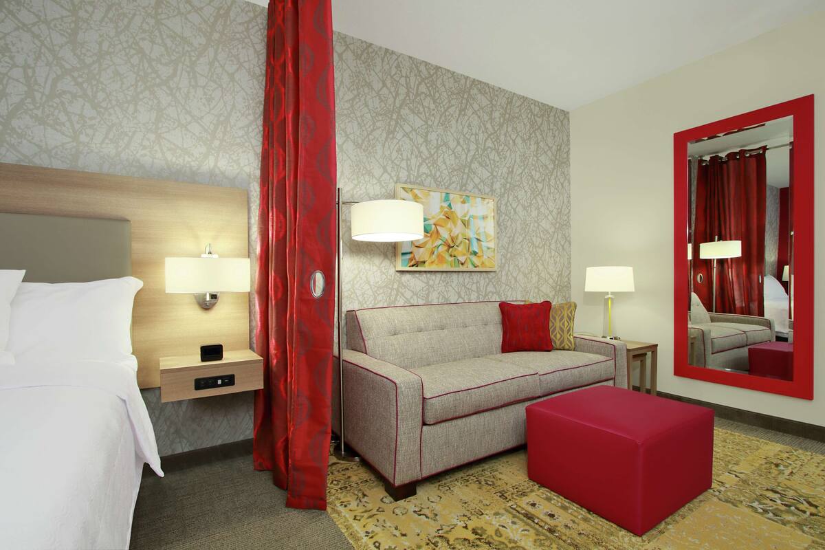 Una camera d'albergo con un letto, una poltrona, un poggiapiedi rosso, uno specchio e un tappeto decorato.