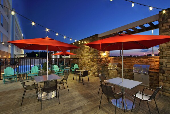 An outdoor patio with tables and chairs, red umbrellas, string lights, and a stone wall with a grill. - Home2 Suites by Hilton Mobile International Airport (Mobile)