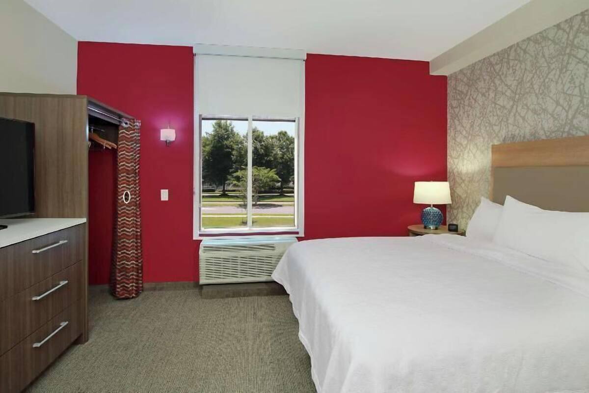 Una camera d'albergo con un letto, una televisione, un armadio, una finestra vista alberi e una lampada.