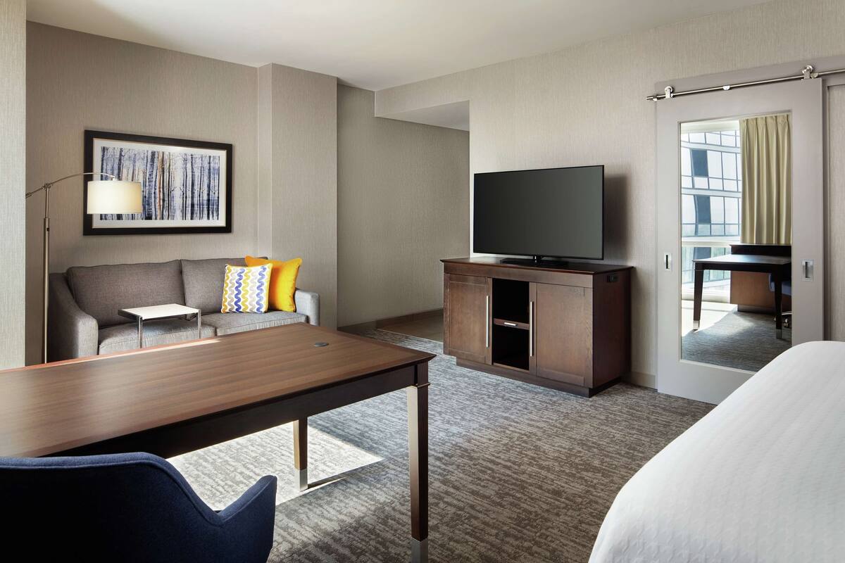 A hotel room with a wooden desk, a sofa with colorful pillows, a flat-screen TV on a wooden cabinet, and a large mirror reflecting the room.