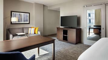 A hotel room with a wooden desk, a sofa with colorful pillows, a flat-screen TV on a wooden cabinet, and a large mirror reflecting the room.