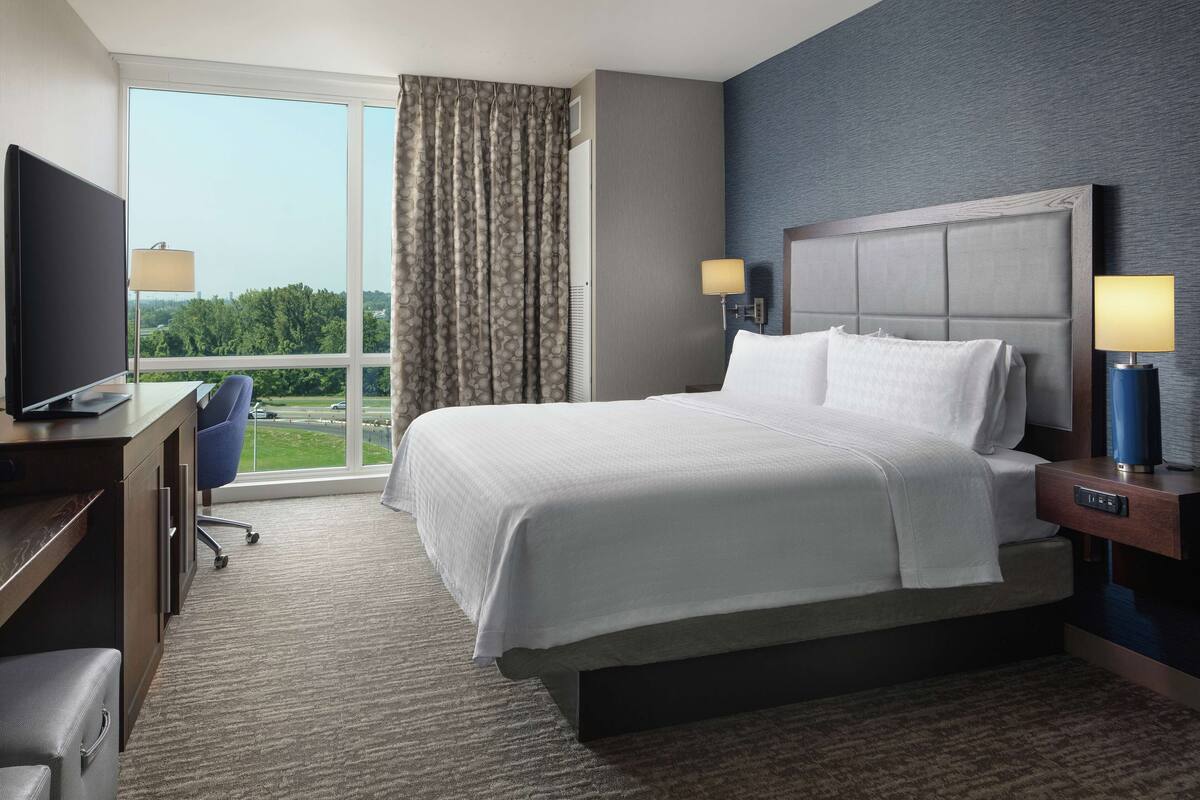 A hotel room with a large bed, a desk with a television, and a view of the outdoors through a large window.