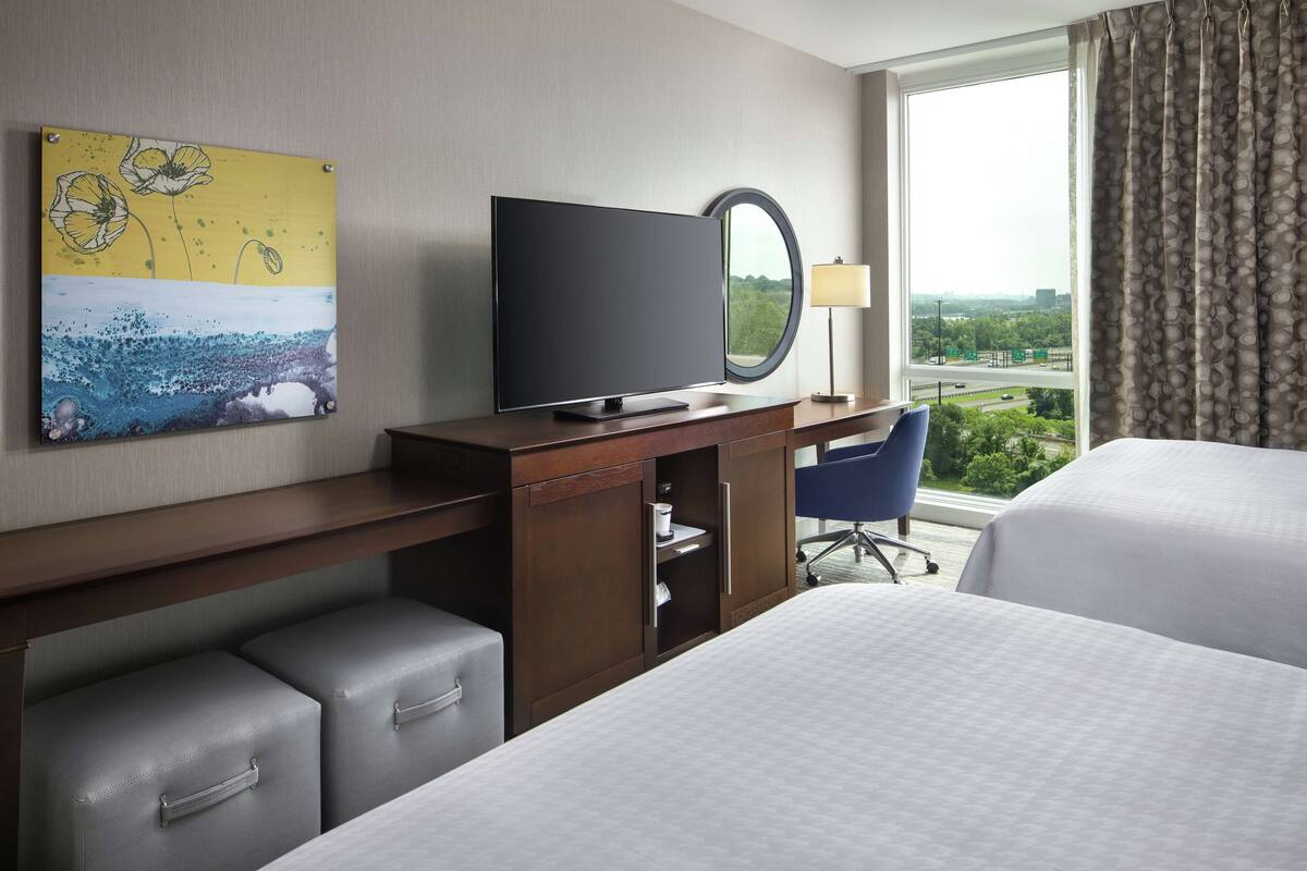 A hotel room with a bed, a desk with a flat-screen TV, a chair, a lamp, and a window with a view of greenery.