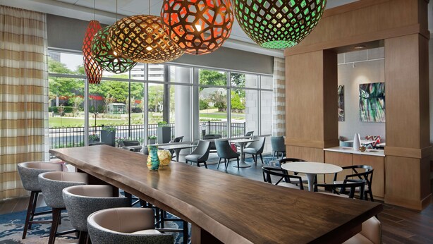 A modern dining area with a long wooden table, multiple chairs, and hanging pendant lights. - Hampton Inn & Suites Teaneck Glenpointe (Teaneck)