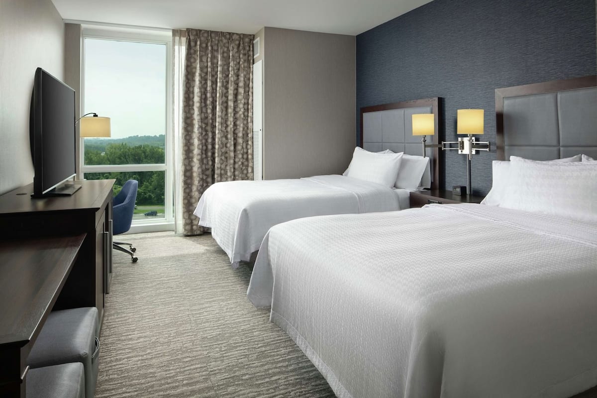 A hotel room with two beds, a desk, a TV, and a view of the outdoors.