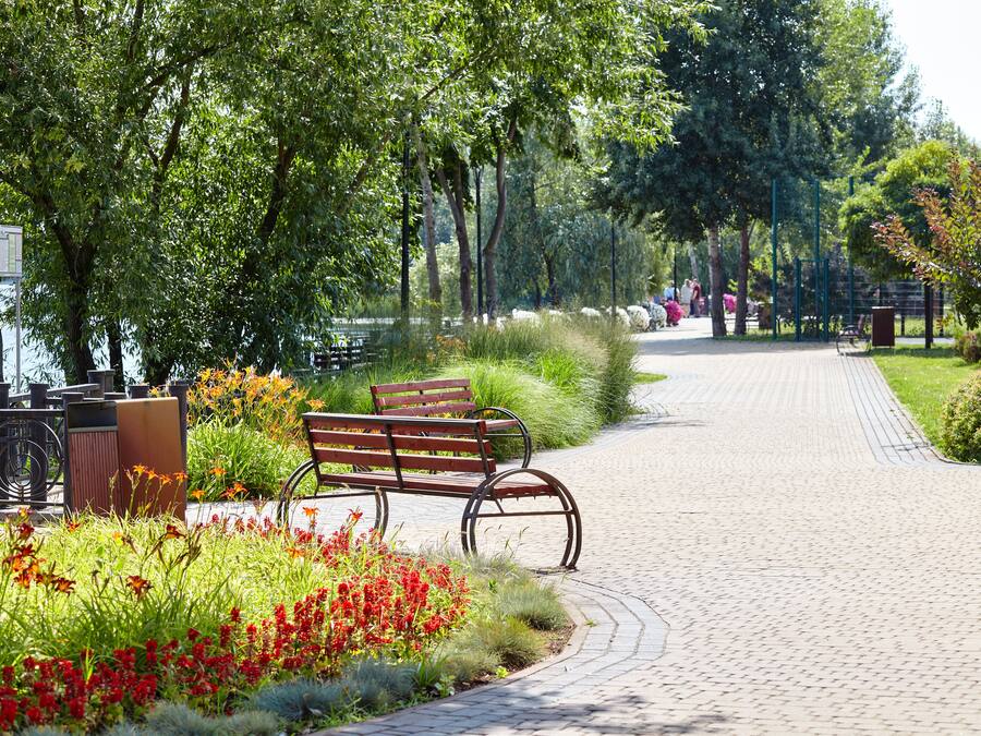 A paved pathway lined with benches and flower beds, surrounded by trees and greenery.