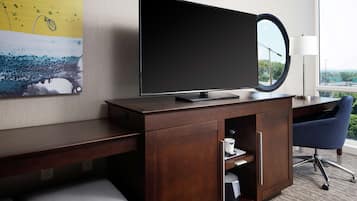 A hotel room with a flat-screen TV mounted on a wooden cabinet, a desk with a chair, a coffee machine, and a window with a view of greenery.