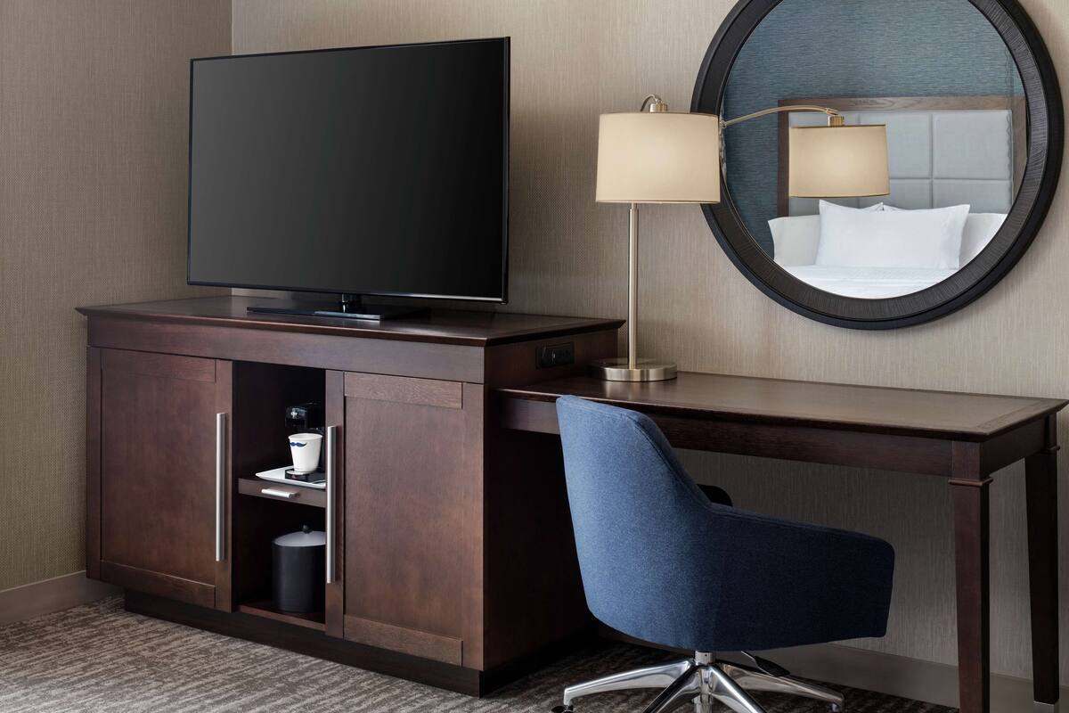 A hotel room with a flat-screen TV, a desk with a chair, a lamp, and a mirror.