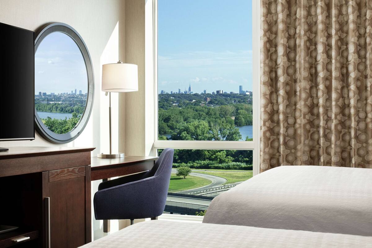 A hotel room with a large window offering a cityscape view, a bed, a desk with a television, and a chair.