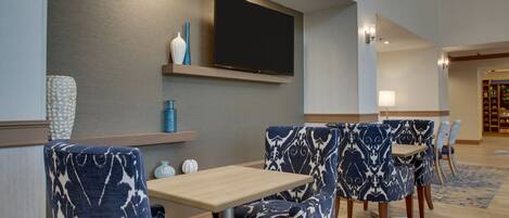 Um hall de hotel moderno com uma TV de tela plana fixada na parede, cadeiras com padrão azul e branco, uma mesa de madeira e uma estante de livros ao fundo.