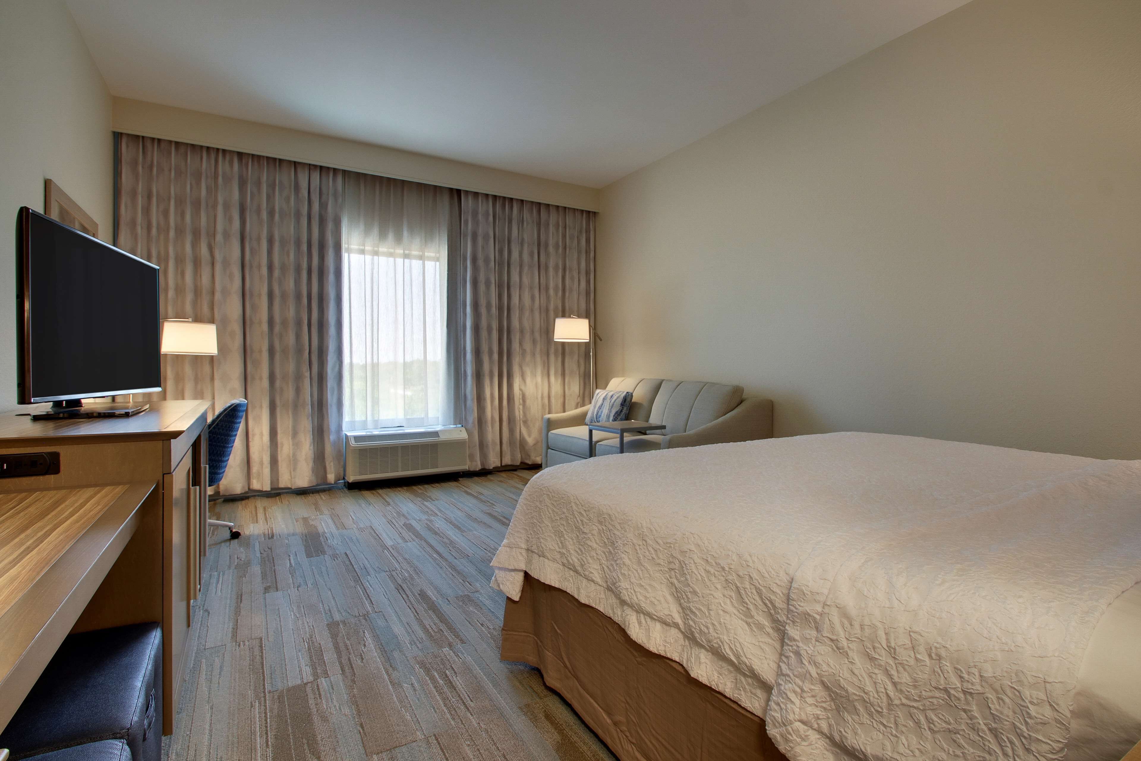 A hotel room with a large bed, a desk with a television, a chair, and a window with curtains.