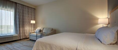 A hotel room with a large bed, a chair, a desk, and a window with curtains.