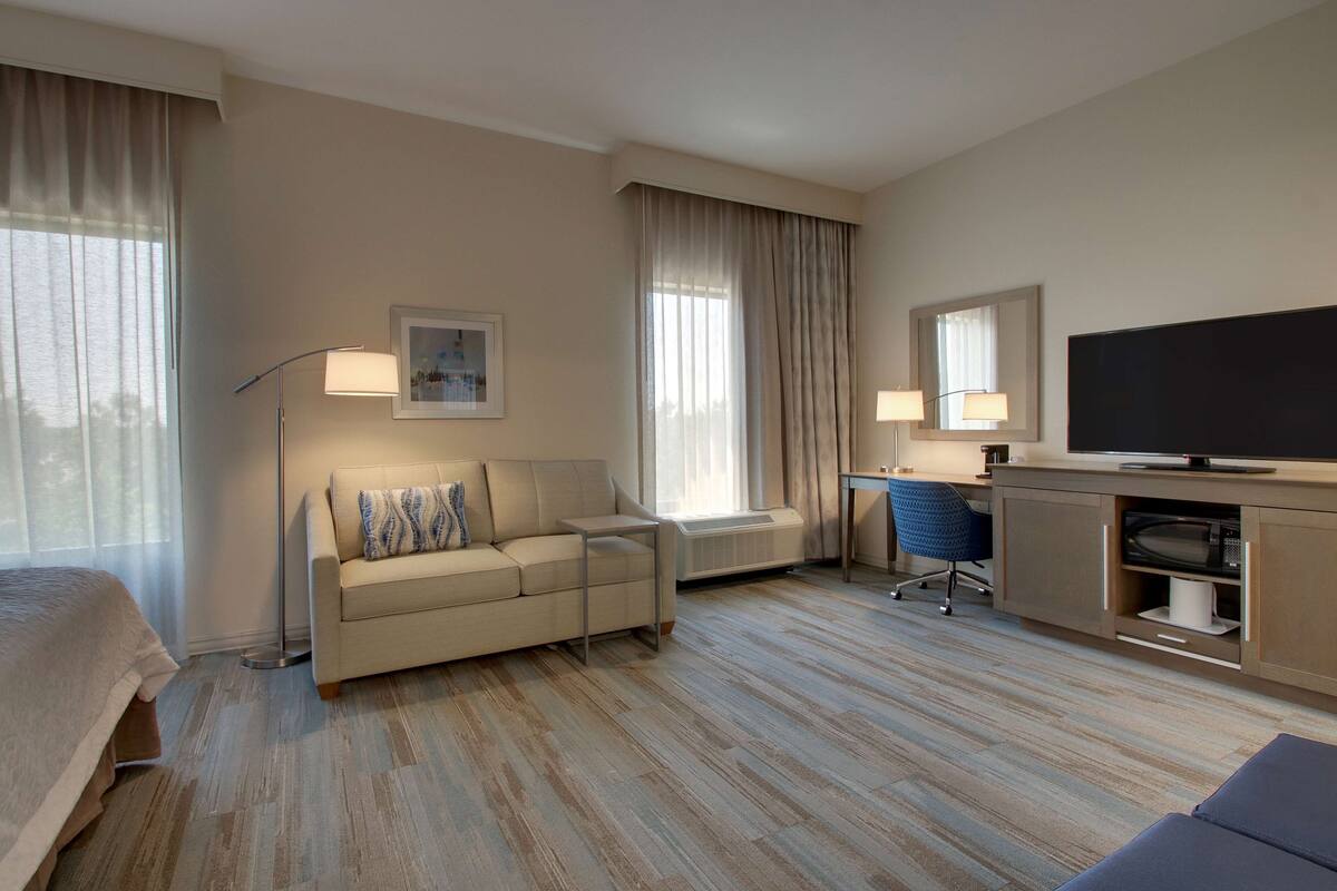 Una habitación de hotel con cama, sofá, escritorio, televisor y ventana con cortinas.