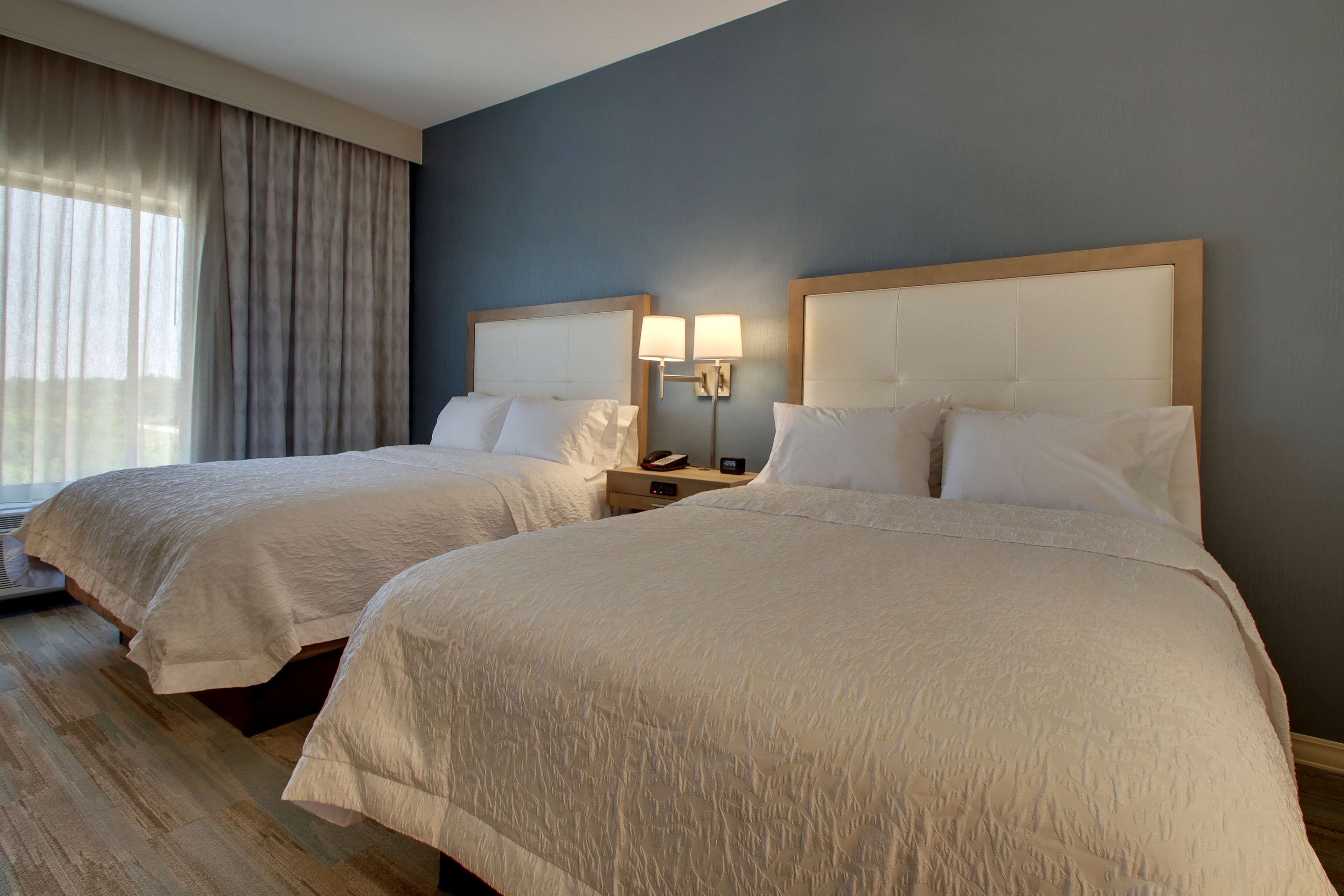 A hotel room with two beds, a nightstand with a lamp, and a large window with curtains.