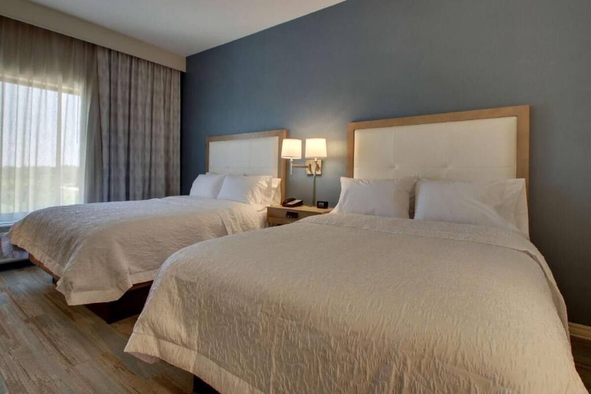 Habitación de hotel con dos camas, un ventanal con cortinas, una mesita de noche con lámpara y una pared azul oscuro.