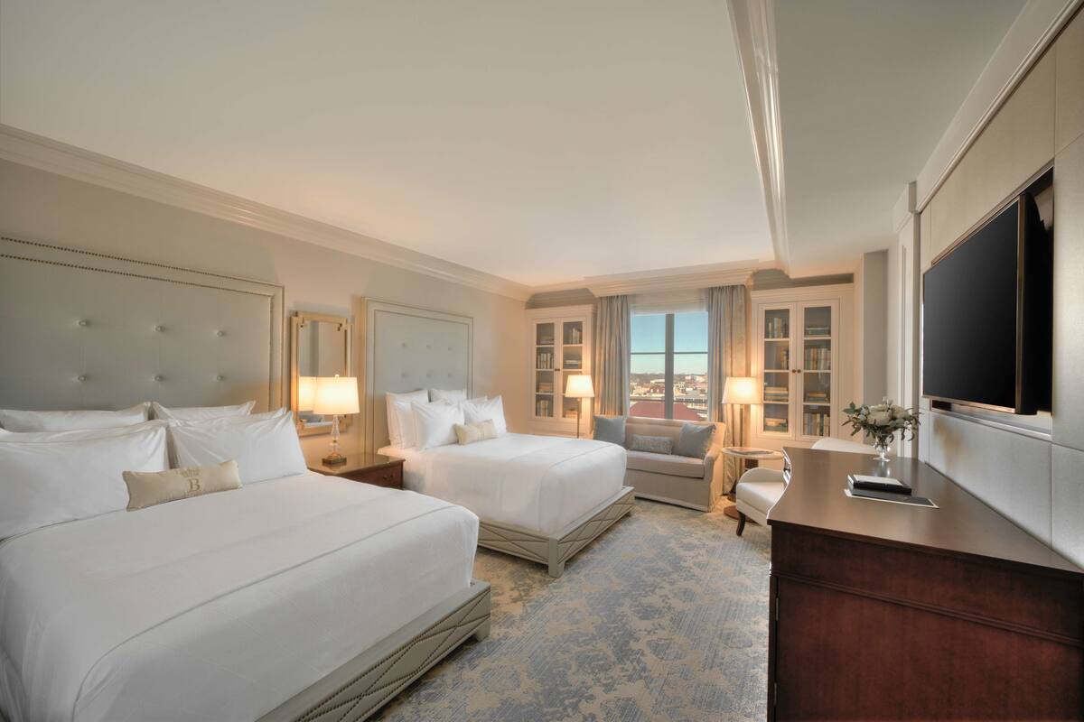 Quarto de hotel com duas camas, um sofá, uma escrivaninha, uma televisão e vista para a cidade.