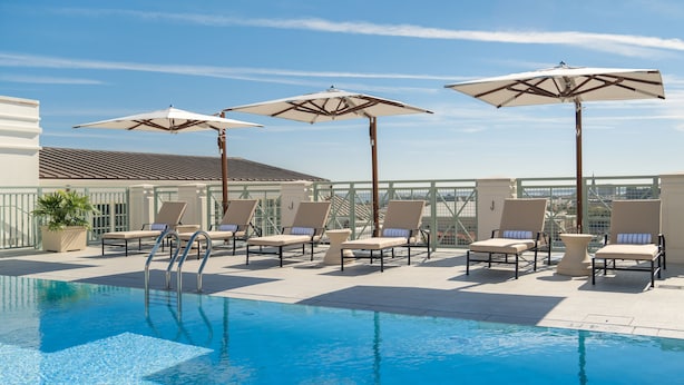 Una piscina en la azotea con sillones de descanso y sombrillas, rodeada por una barandilla de cristal y un edificio con techo de tejas.