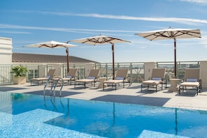 A rooftop pool with lounge chairs and umbrellas, surrounded by a glass railing and a building with a tiled roof. - Hotel Bennett (Charleston)