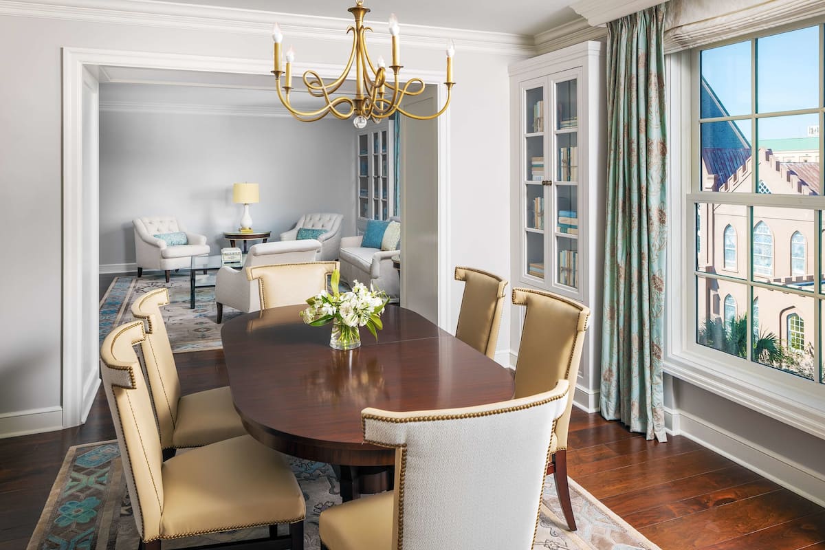 Uma sala de jantar com uma mesa redonda, seis cadeiras, um candelabro e vista para edifícios pela janela.