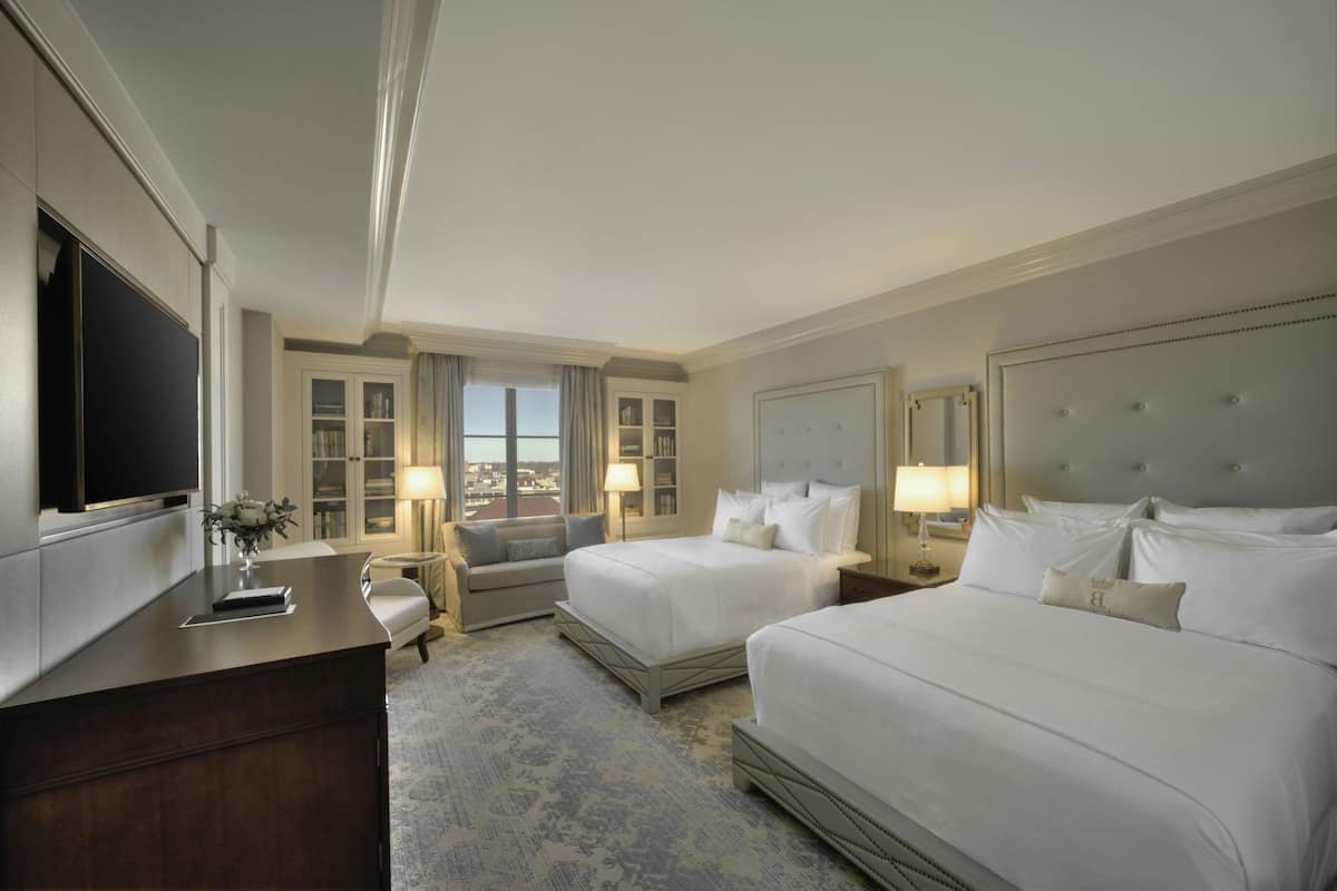 A hotel room with two beds, a desk, a TV, and a view of the city.