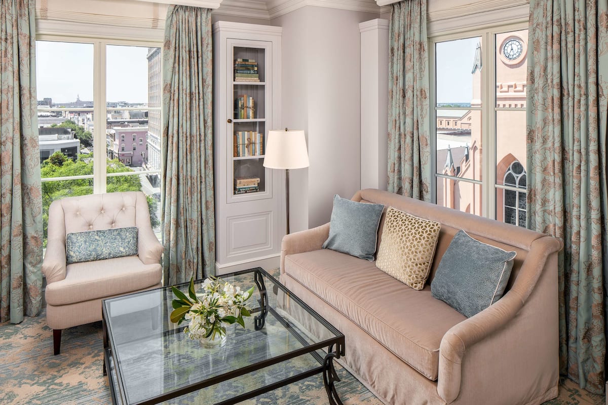 Quarto com sofá bege, poltrona estofada, mesa de centro de vidro e um arranjo floral, além de uma estante de livros.