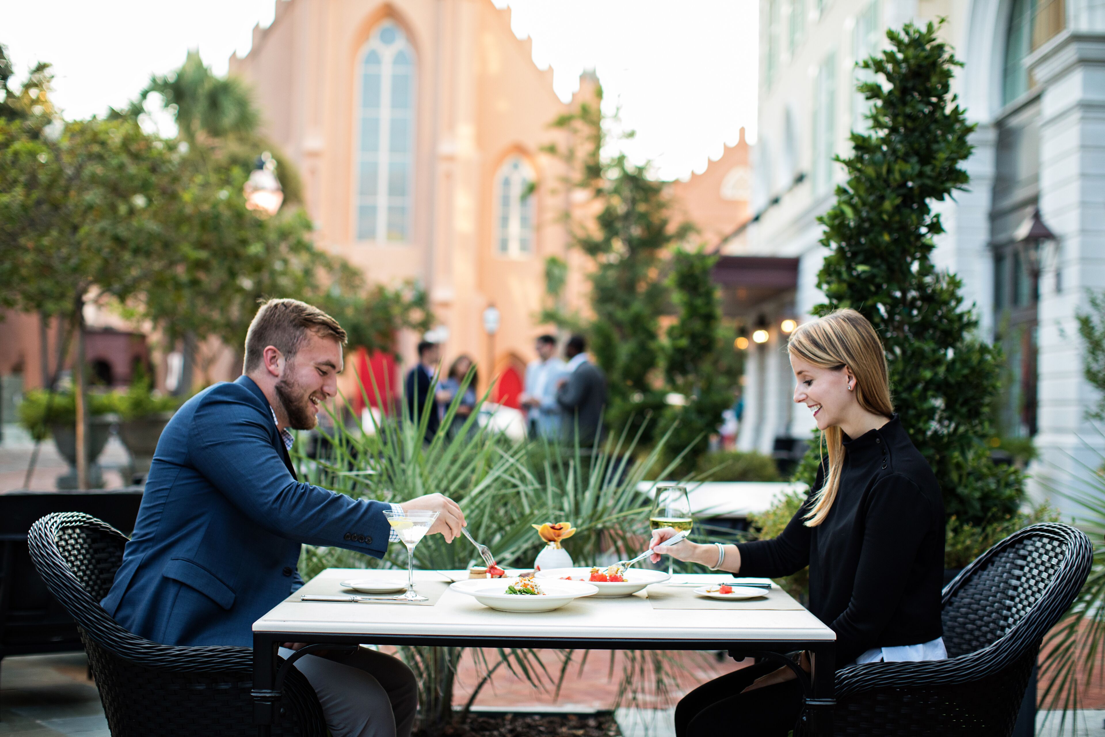 A couple dining at an outdoor table with a view of a church and other buildings in the background.