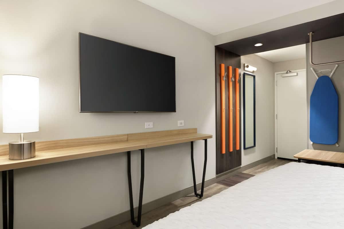 A modern hotel room with a large flat-screen TV mounted on the wall, a wooden console table, a bed with a white comforter, and a blue ironing board.
