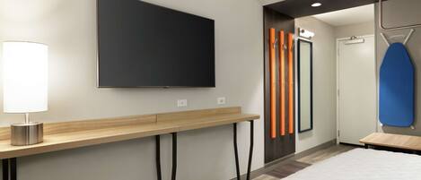 A modern hotel room with a large flat-screen TV mounted on the wall, a wooden console table, a bed with a white comforter, and a blue ironing board.