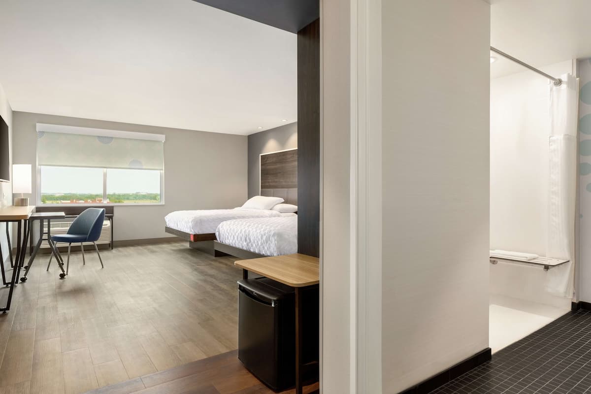 A modern hotel room with a wooden floor, a desk with a chair, a bed with white bedding, and a window with blinds.