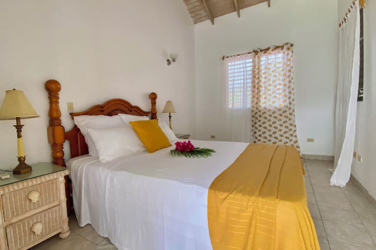 Una cama bien hecha con ropa blanca y un edredón amarillo, una mesita de noche de madera con una lámpara, y una mesita auxiliar de mimbre con toma de corriente.