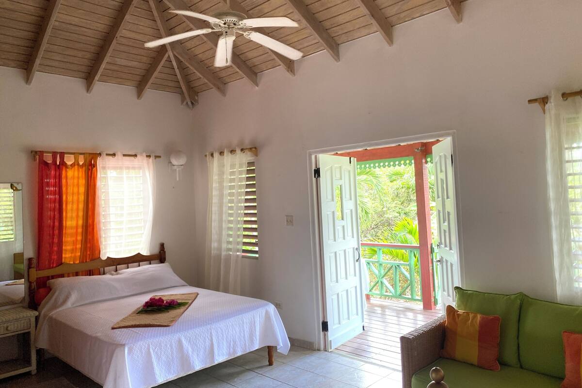 Un dormitorio con cama, ventilador de techo, ventana con cortinas y puerta abierta que da a un balcón.