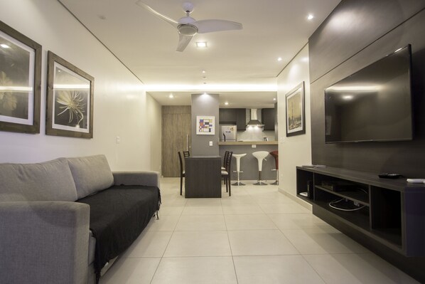 A modern living room with a grey sofa, a dining area with a table and chairs, a kitchen in the background, and a mounted TV.