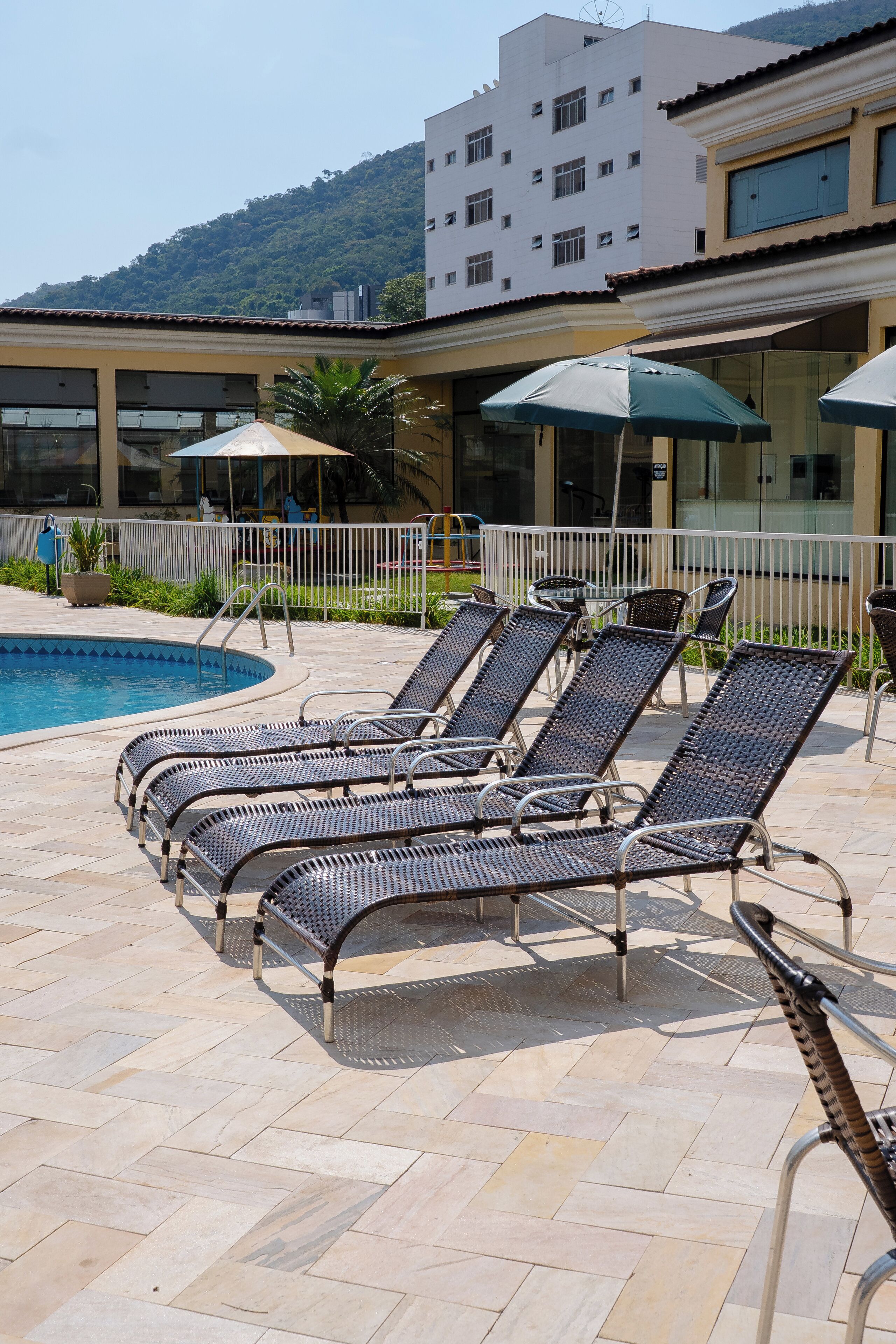 Zona junto a la piscina con sillones y sombrillas, una alberca y un edificio con ventanas amplias y un balcón.