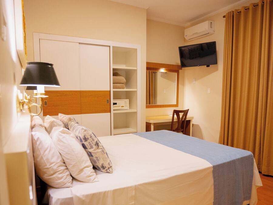 A hotel room with a bed, bedside table with a telephone, a desk with a chair, a television mounted on the wall, and a wardrobe.