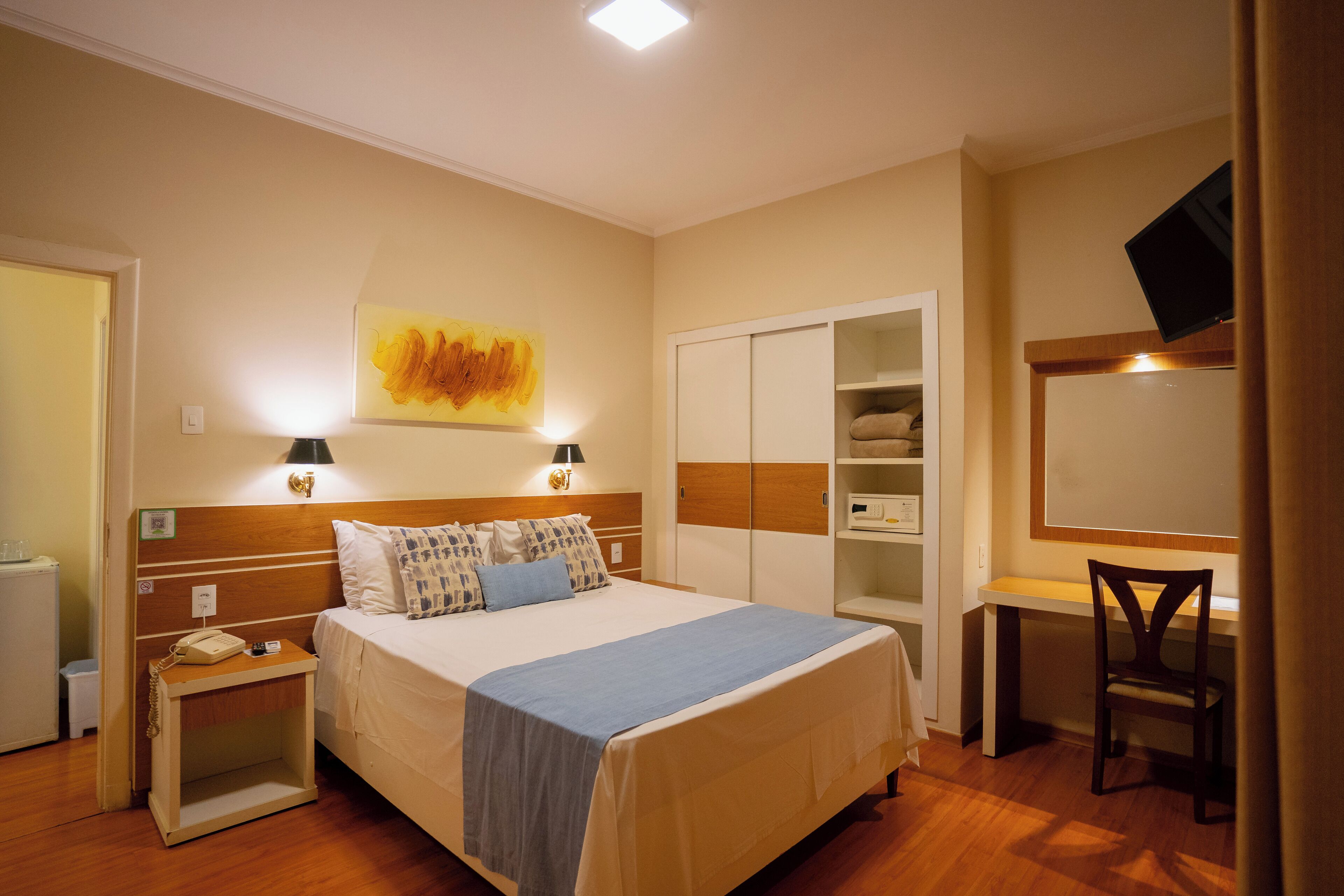 Habitación de hotel con cama, mesita de noche, lámpara, armario, escritorio, silla y televisor montado en la pared.