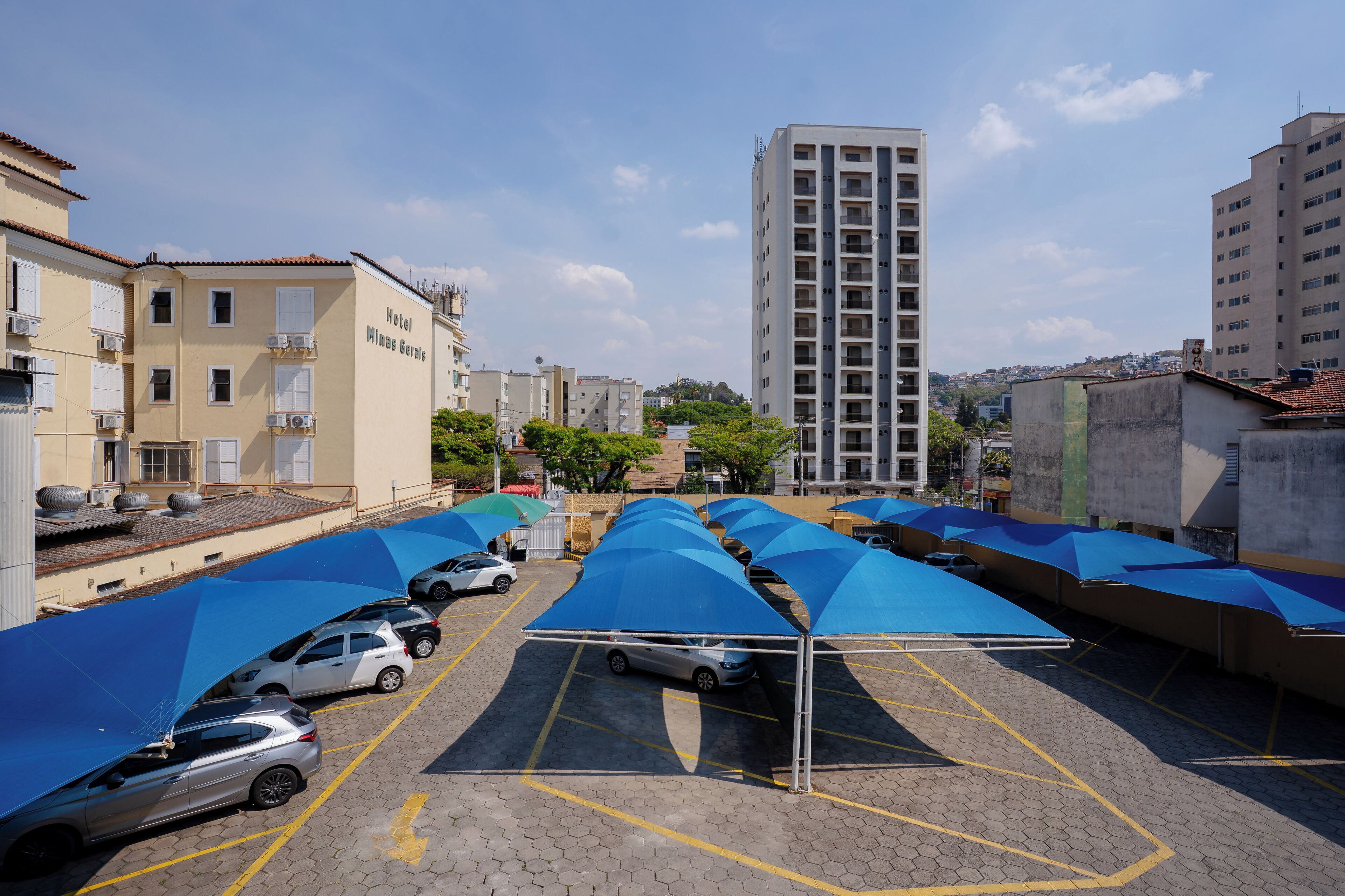 Un área de estacionamiento con toldos azules, un edificio con la leyenda "Hotel Minas Gerais" y un edificio residencial alto al fondo.