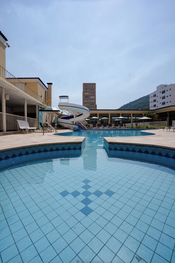 Una piscina con sillones de descanso y sombrillas, rodeada de edificios y un paisaje montañoso.