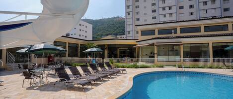 Zona de piscina con sillones de descanso y sombrillas, rodeada por un edificio de varios pisos y vista a una colina.