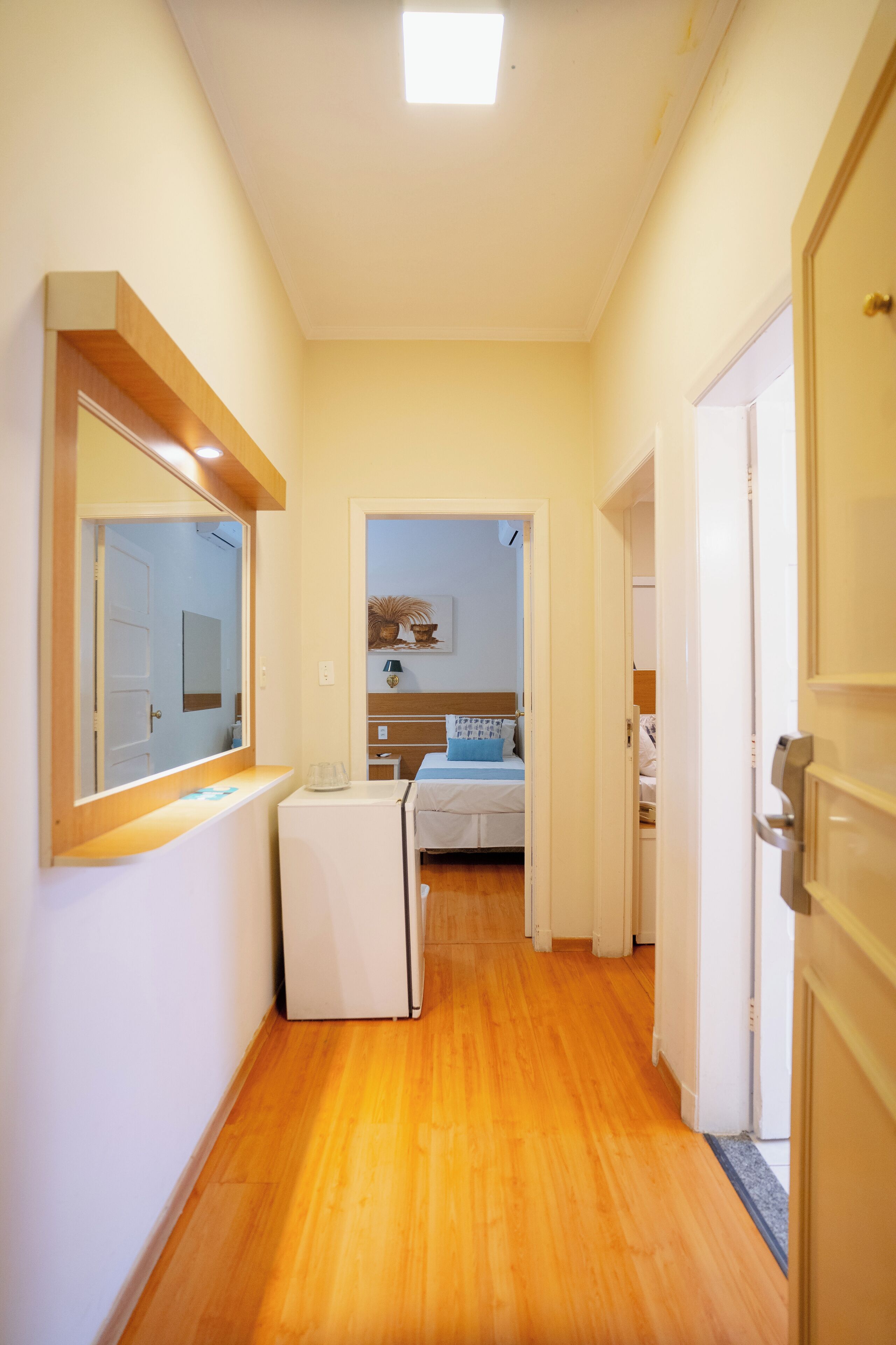Un pasillo estrecho con suelo de madera que conduce a un dormitorio con una cama y una mesita de noche. Se observa un electrodoméstico blanco pequeño a la izquierda.