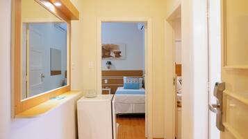 Un pasillo estrecho con suelo de madera que conduce a un dormitorio con una cama y una mesita de noche. Se observa un electrodoméstico blanco pequeño a la izquierda.