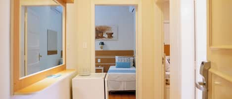 Un pasillo estrecho con suelo de madera que conduce a un dormitorio con una cama y una mesita de noche. Se observa un electrodoméstico blanco pequeño a la izquierda.