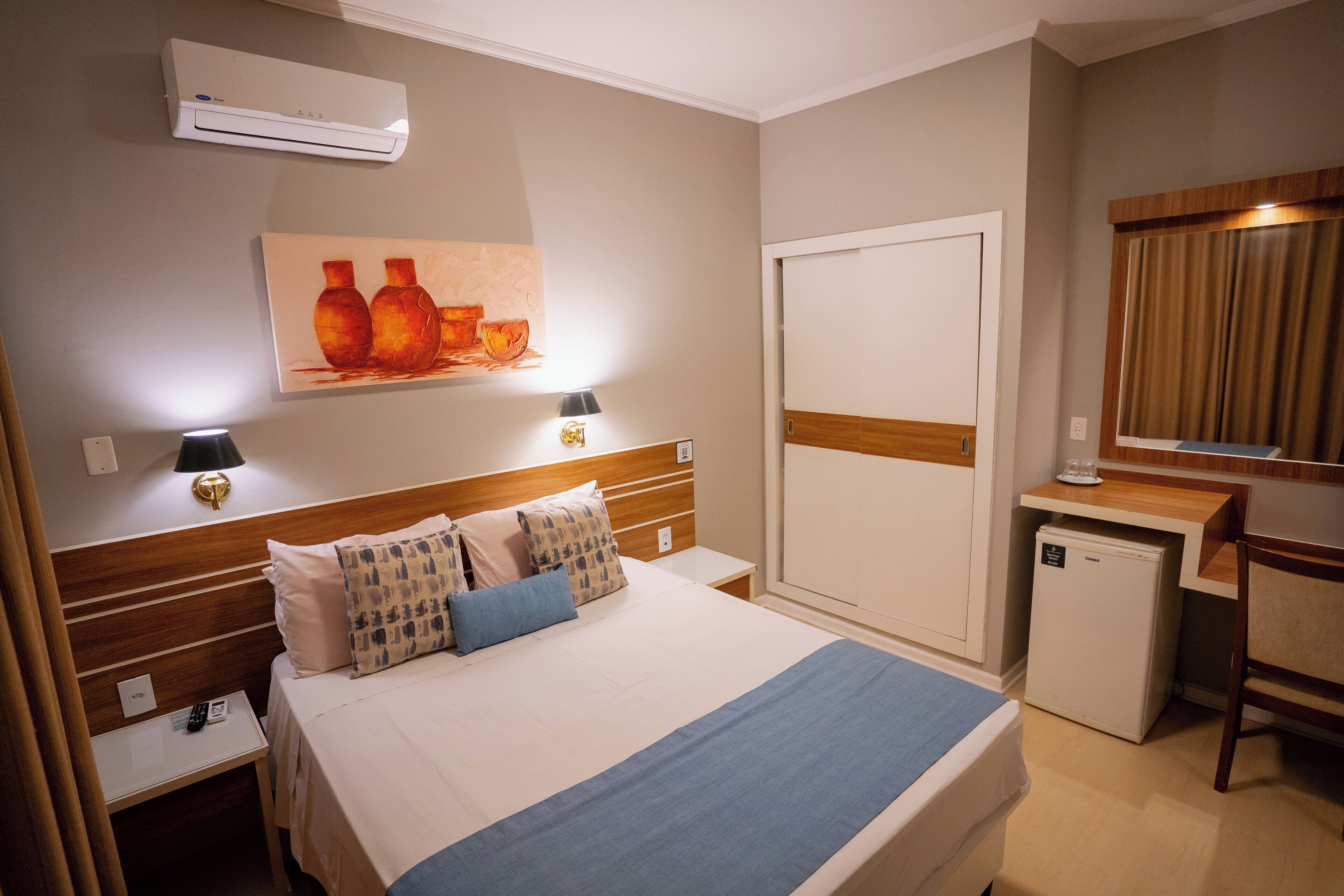 A hotel room with a bed, bedside tables, a desk, a refrigerator, and a wall-mounted air conditioner.