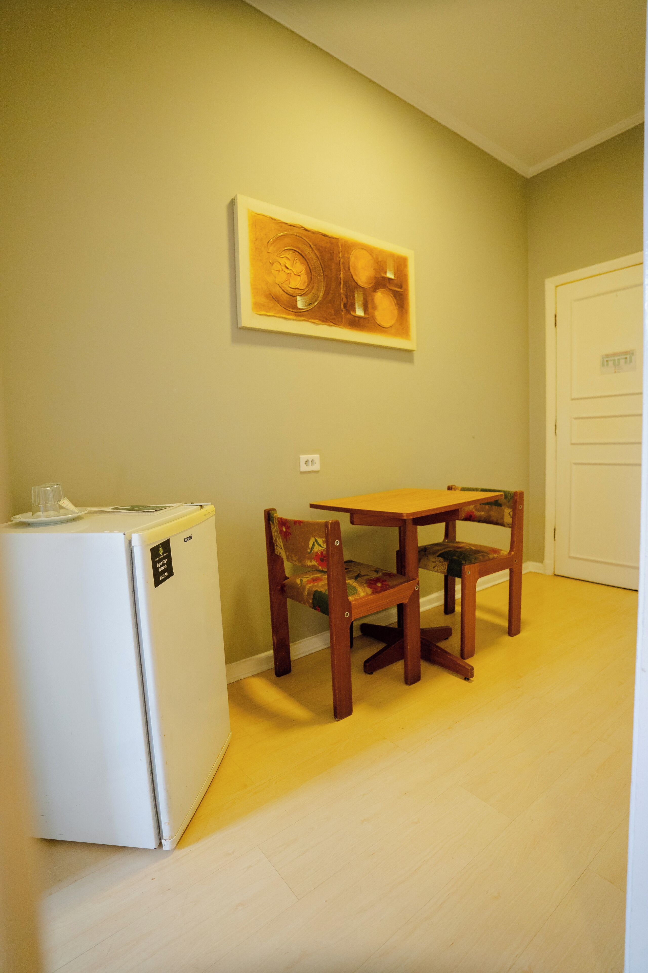 Een kleine keuken met een witte koelkast, een houten tafel en een stoel met bloemenmotief.