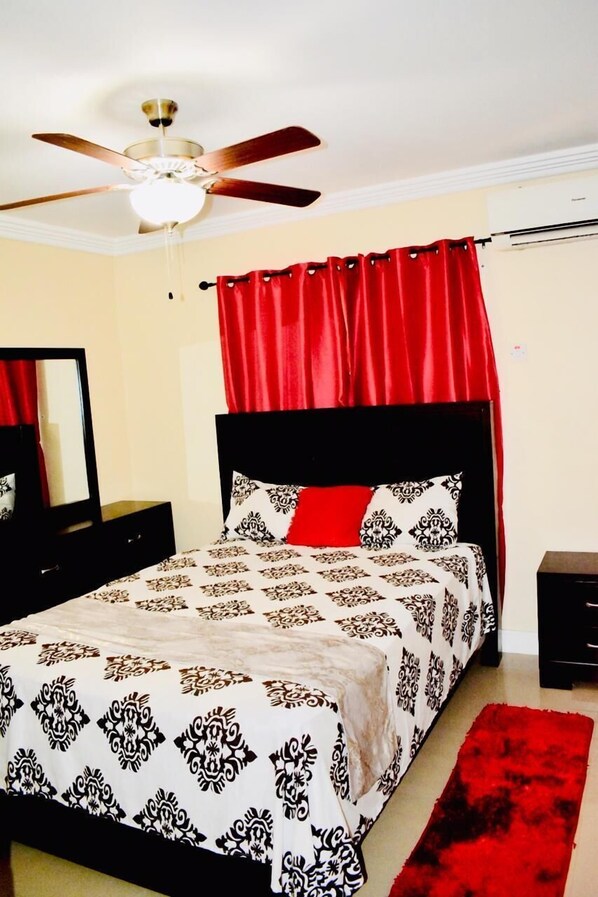 A bedroom with a bed, a ceiling fan, a red curtain, a black nightstand, and a mirror. - Kingston Guest Apartment at El Gabriel (Kingston)