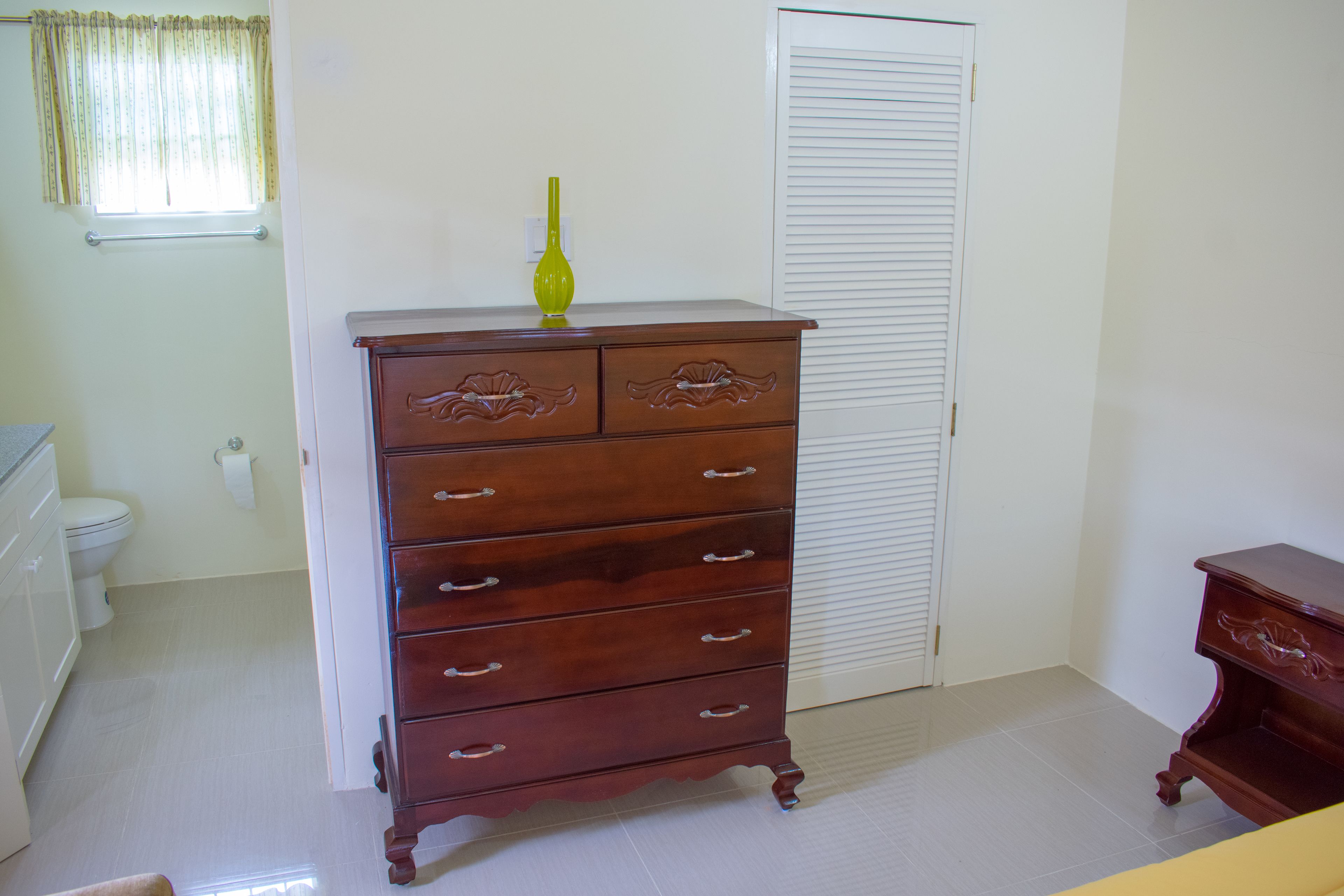 A bathroom with a wooden dresser, a toilet, and a window with curtains.
