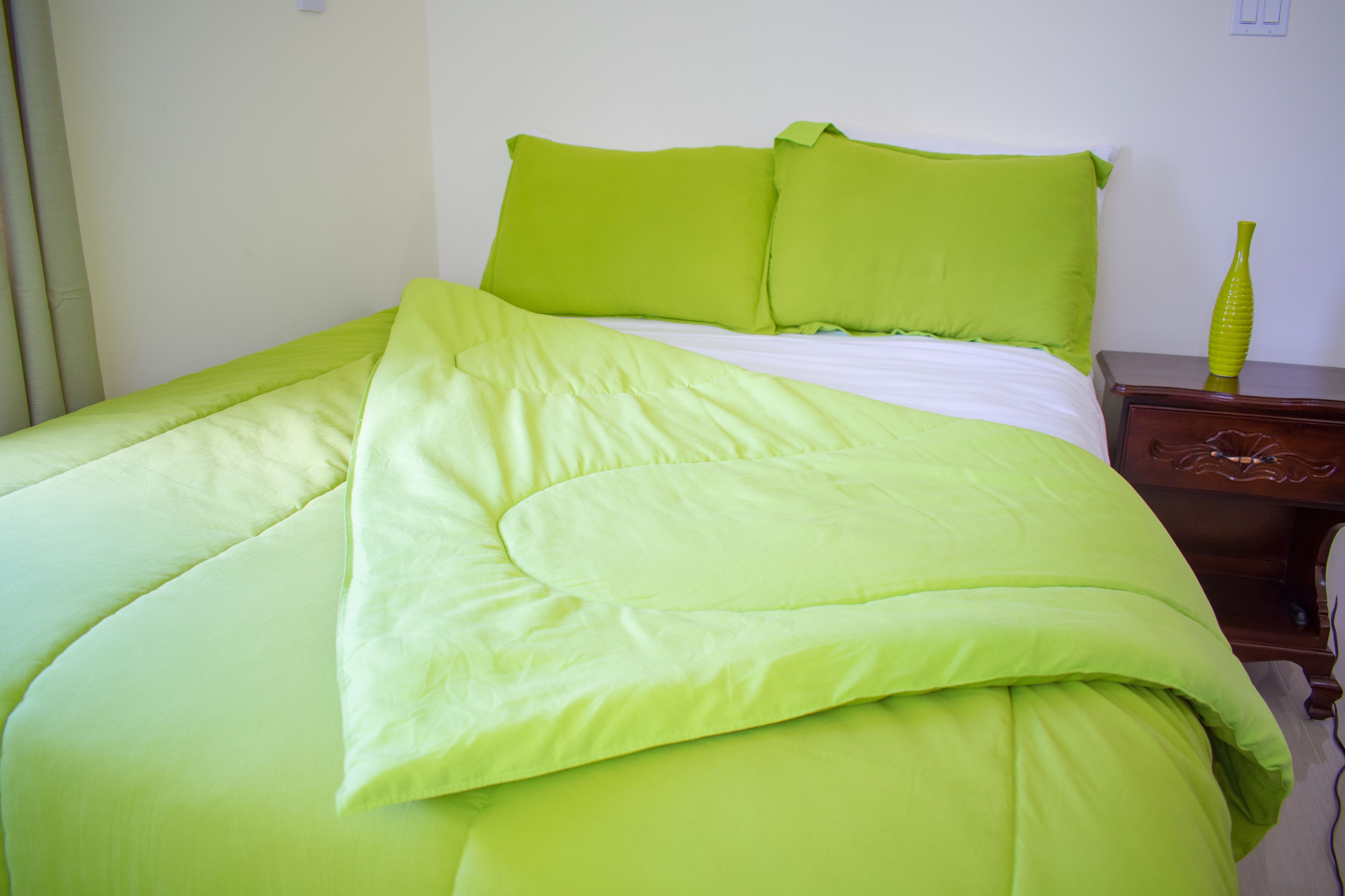 Una cama con ropa de cama verde y una mesita de noche de madera con un florero verde.